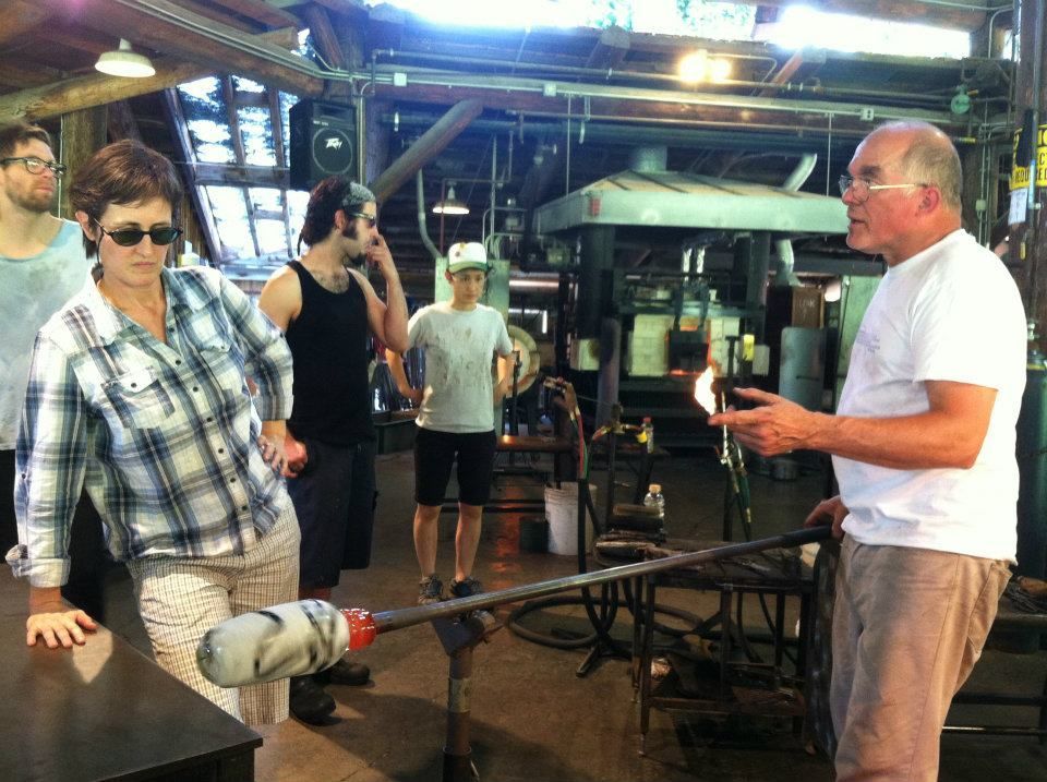 Glass Artist, John de Wit, demonstrates with molten glass, surrounded by onlookers in a workshop.