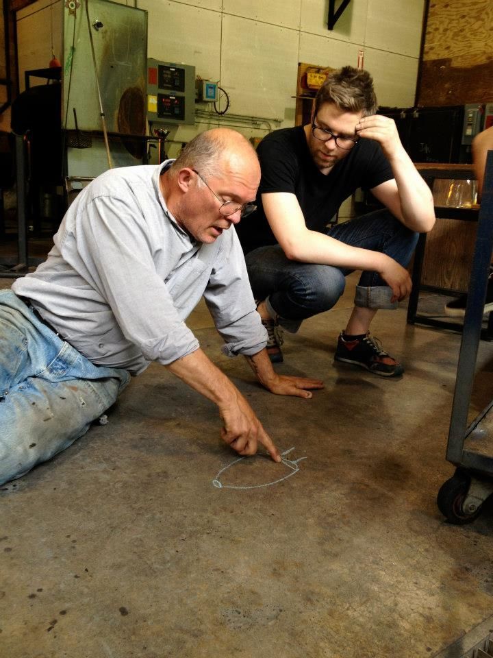 Glass Artist, John de Wit, is pointing at a chalk drawing on the floor as another man watches in a workshop.