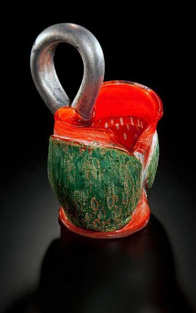 Glass sculpture of a red and green abstract form with a silver handle, set against a black background.