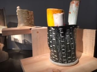 Ceramic vase with tall, colorful cylinders. Black basketweave design on the vase, set on a wooden shelf.