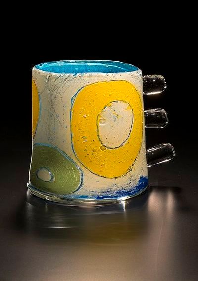 Glass mug with yellow and blue circles on a white surface, with three clear protrusions on the side.