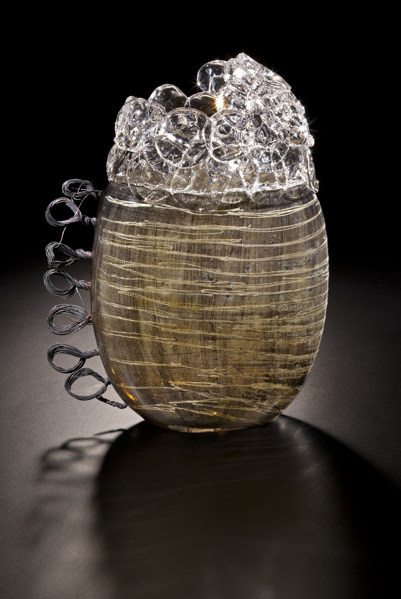 Sculptural vessel with a wire spiral, textured glass body, and bubbly top against a dark backdrop.