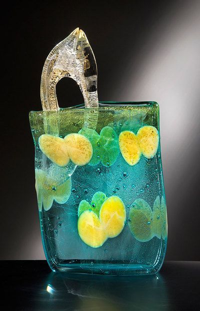 Glass sculpture: teal vase with yellow and green flower-like shapes and a clear handle on a dark background.