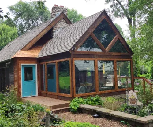 Brown-shingled cottage with teal door and large windows; sunroom with triangular window, set among trees.