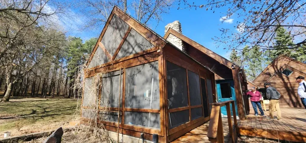 A rustic cabin with a screened porch is surrounded by trees; people stand nearby.
