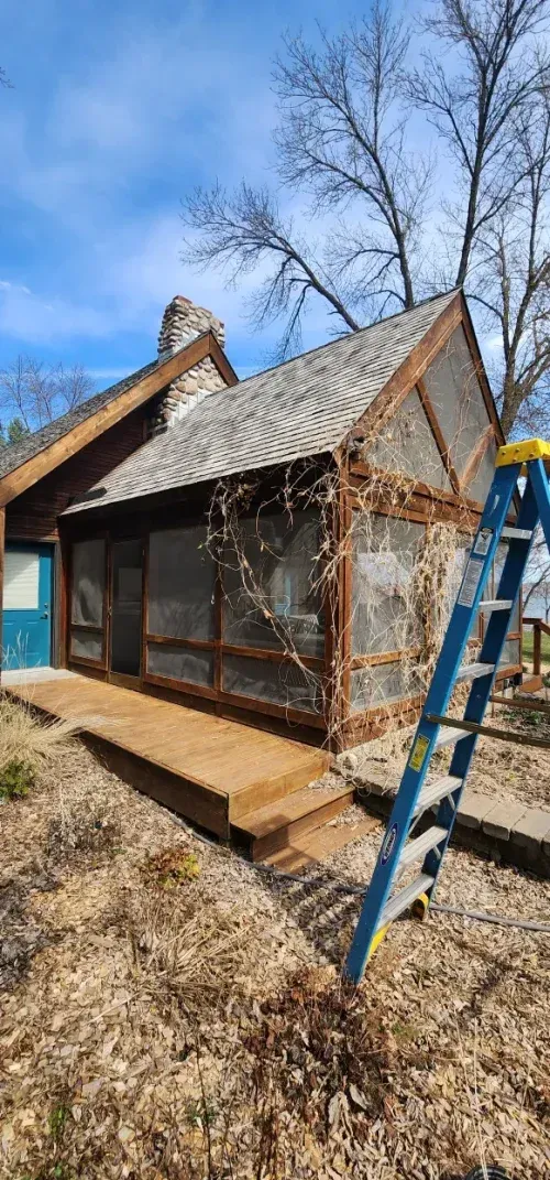A wooden screened-in porch and wooden deck with a blue ladder, in a yard with brown mulch, trees and a chimney.