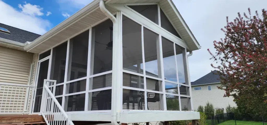 A screened-in porch with white trim and screens, attached to a house with a blue sky background.