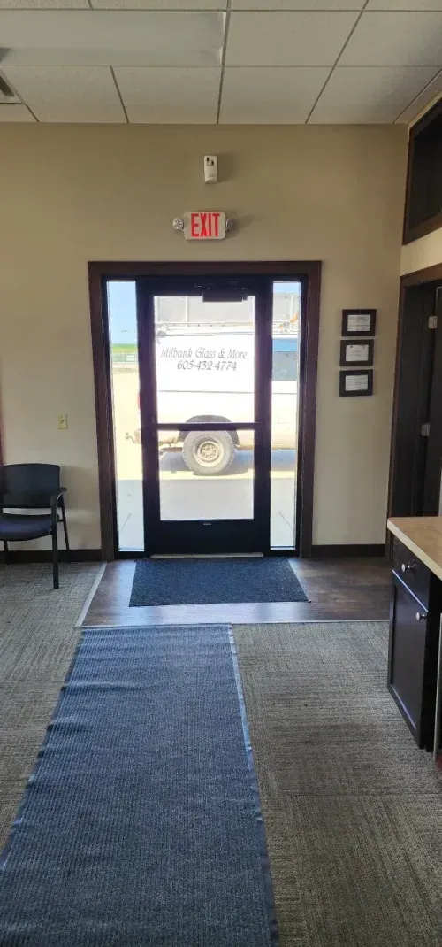 Interior view of a door with an exit sign above. A blue rug leads to the door, which has a vehicle parked on the other side.