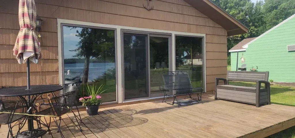 Lakeside home with patio and furniture. Brown siding, large windows, and wooden deck. Umbrella and chairs visible.