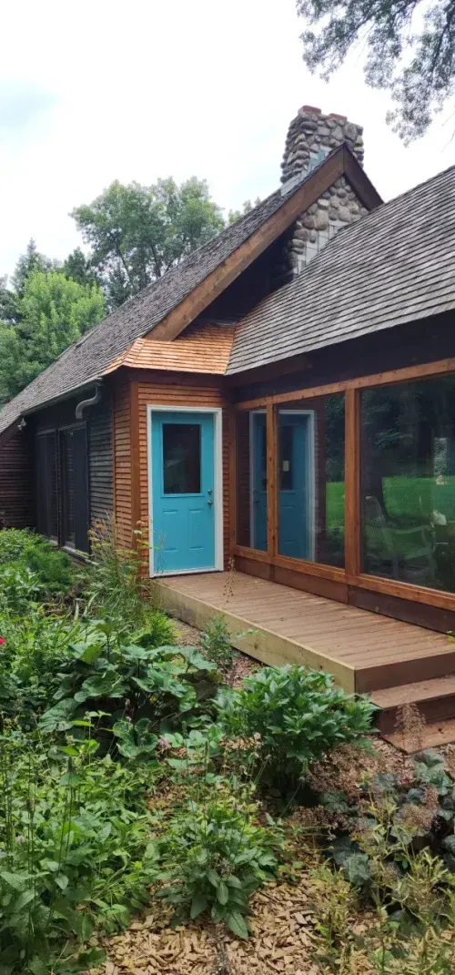 Cabin with a turquoise door, wooden deck, and large windows; surrounded by greenery.