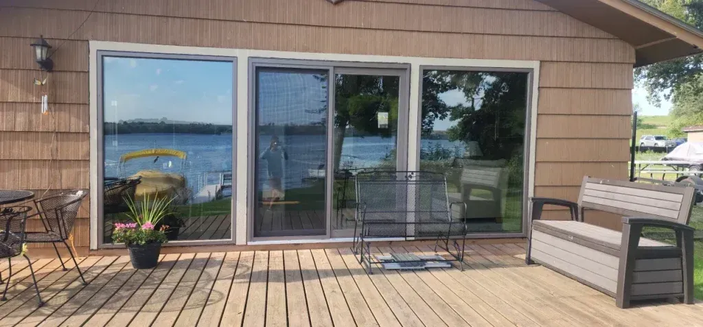A lakeside house with a wooden deck, windows, and outdoor furniture.