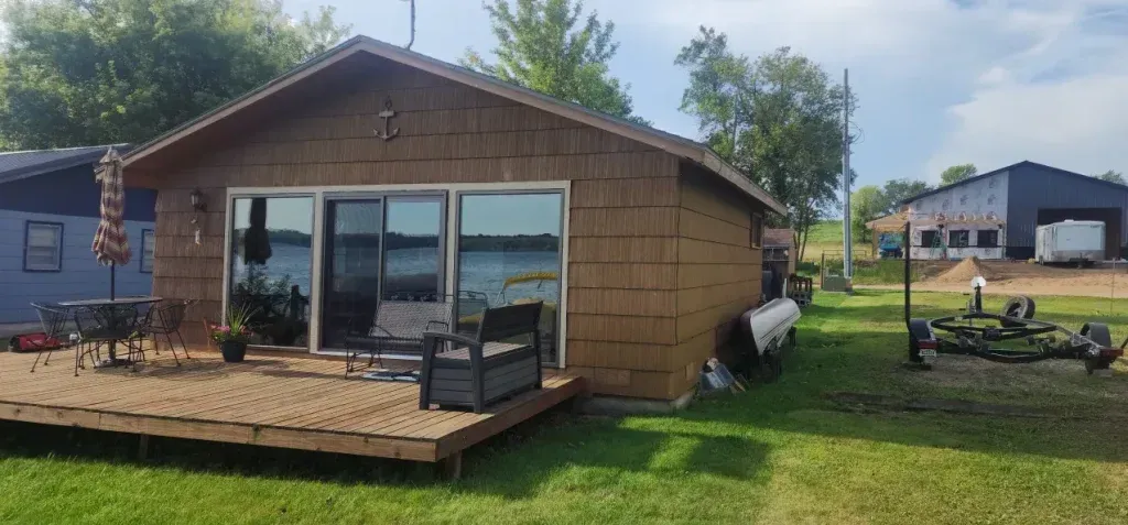 Brown lake house with a deck, glass sliding doors, and a lawn. A boat trailer sits on the grass.