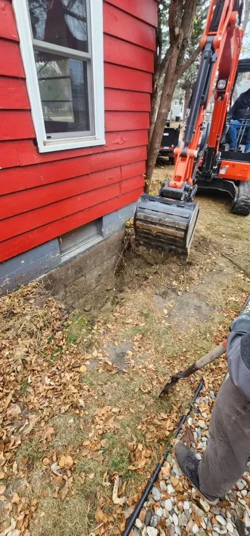 An excavator digs near the foundation of a red house. A person with a shovel is present.