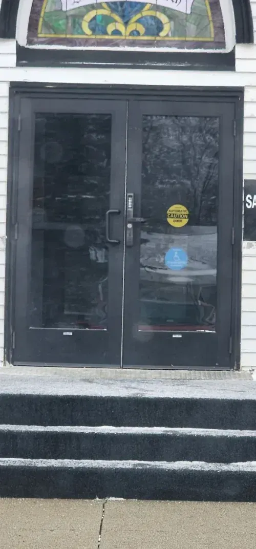 Black double doors with a yellow accessibility sticker, leading into a white building, with three steps leading up to the doors.