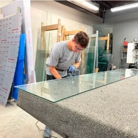 A young man in gray shirt grinding the edge of a large glass pane in a workshop.