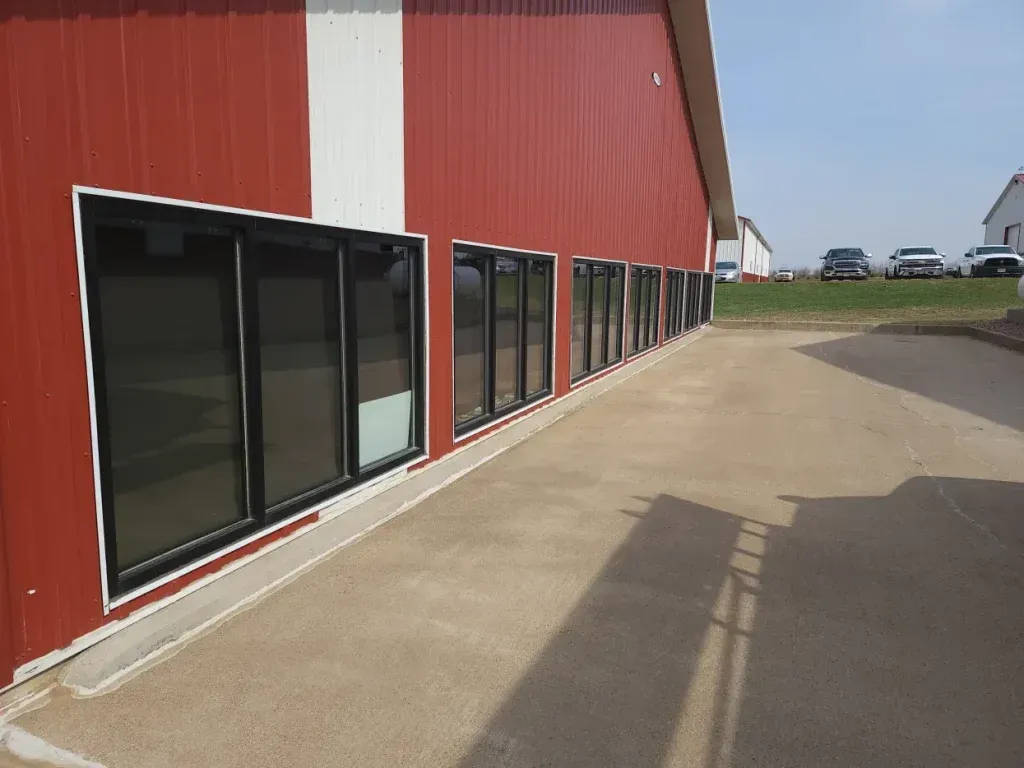 Red building with black-framed windows along a concrete walkway. Cars parked in the background on a sunny day.