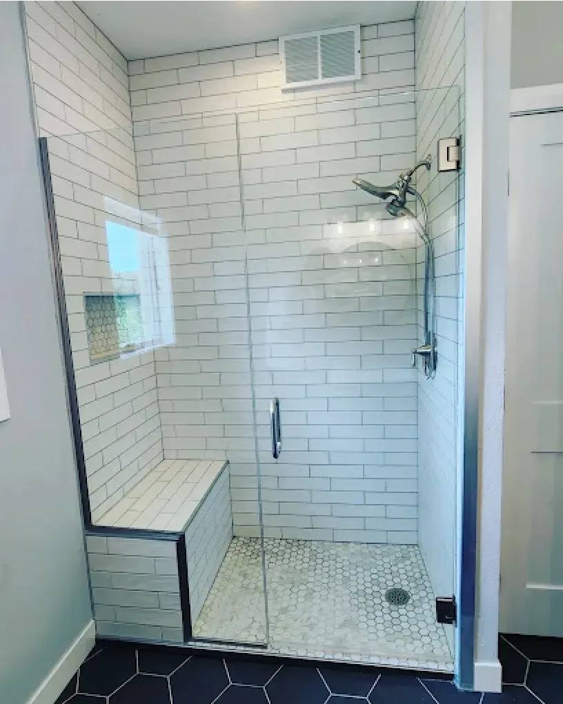 Modern white tiled shower with glass door and built-in bench, black hexagonal floor tiles, and gray walls.