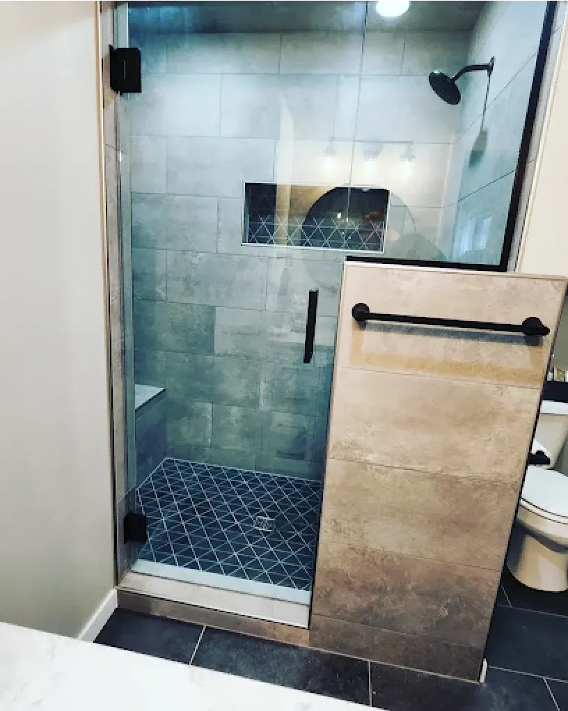 Shower with glass door, gray tile walls, black showerhead, and dark blue patterned floor.