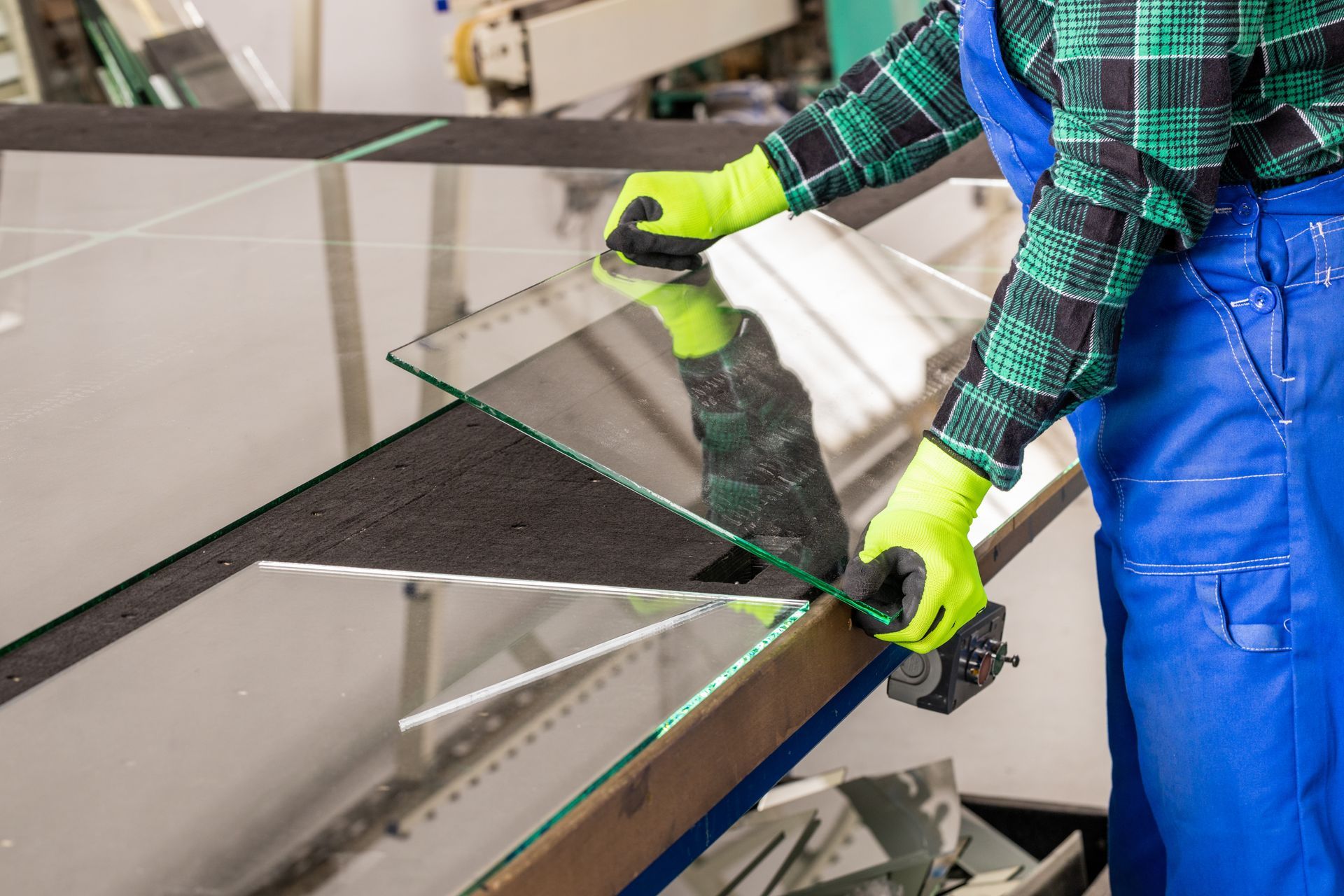 A worker in blue overalls and green gloves carefully handles a large glass sheet, likely in a factory.