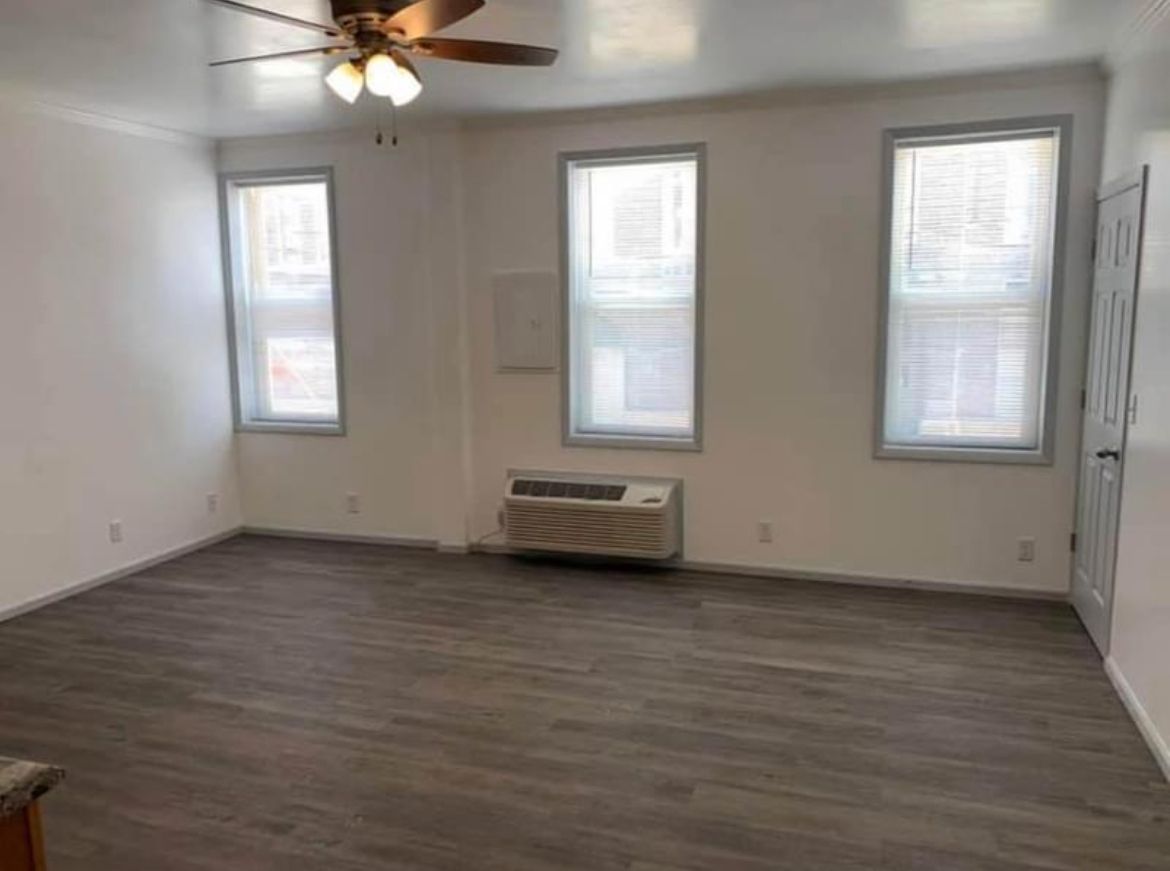 An empty living room with a ceiling fan and three windows.