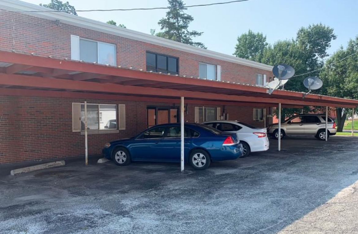 A row of cars are parked under a canopy in front of a brick building.