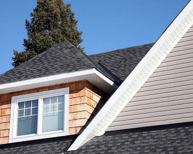 Close-up of house with gray roof, brown cedar siding, white trim, and a blue sky.