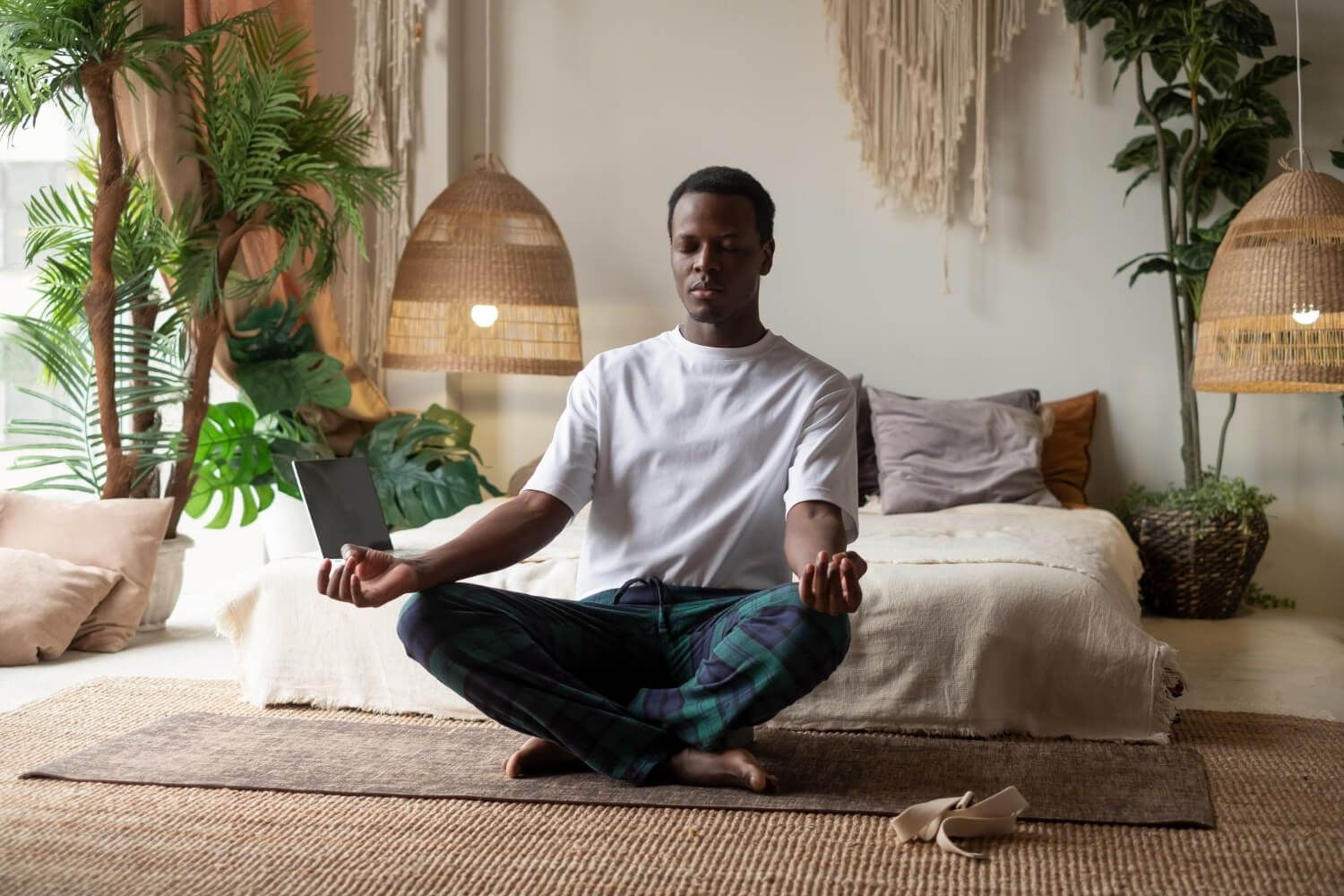 Young Black man in white t-shirt sitting cross-legged on bed in peaceful living room practicing medi