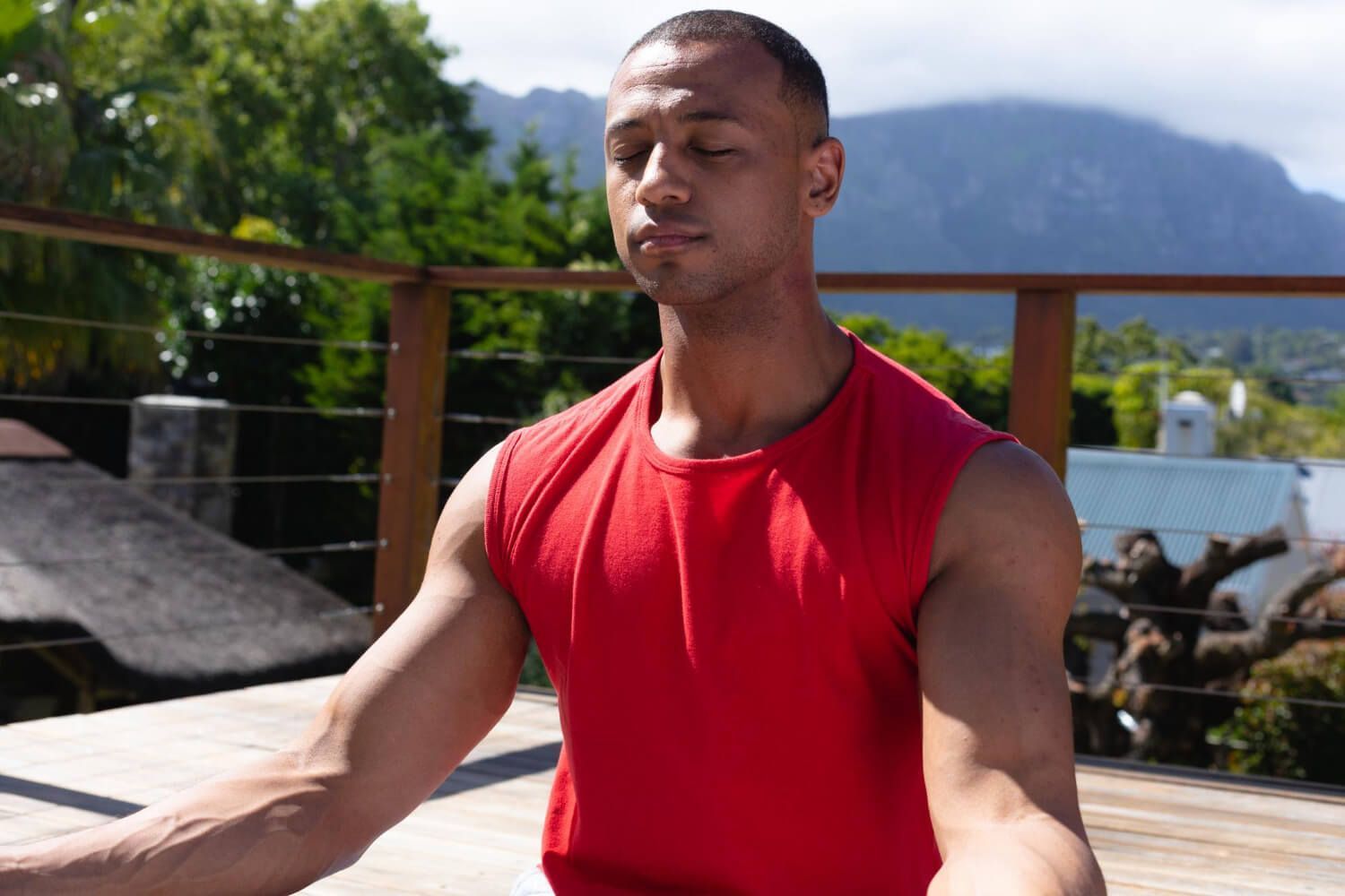 African American man in red shirt practicing breathing exercise outdoors with eyes closed, mountains and greenery in background