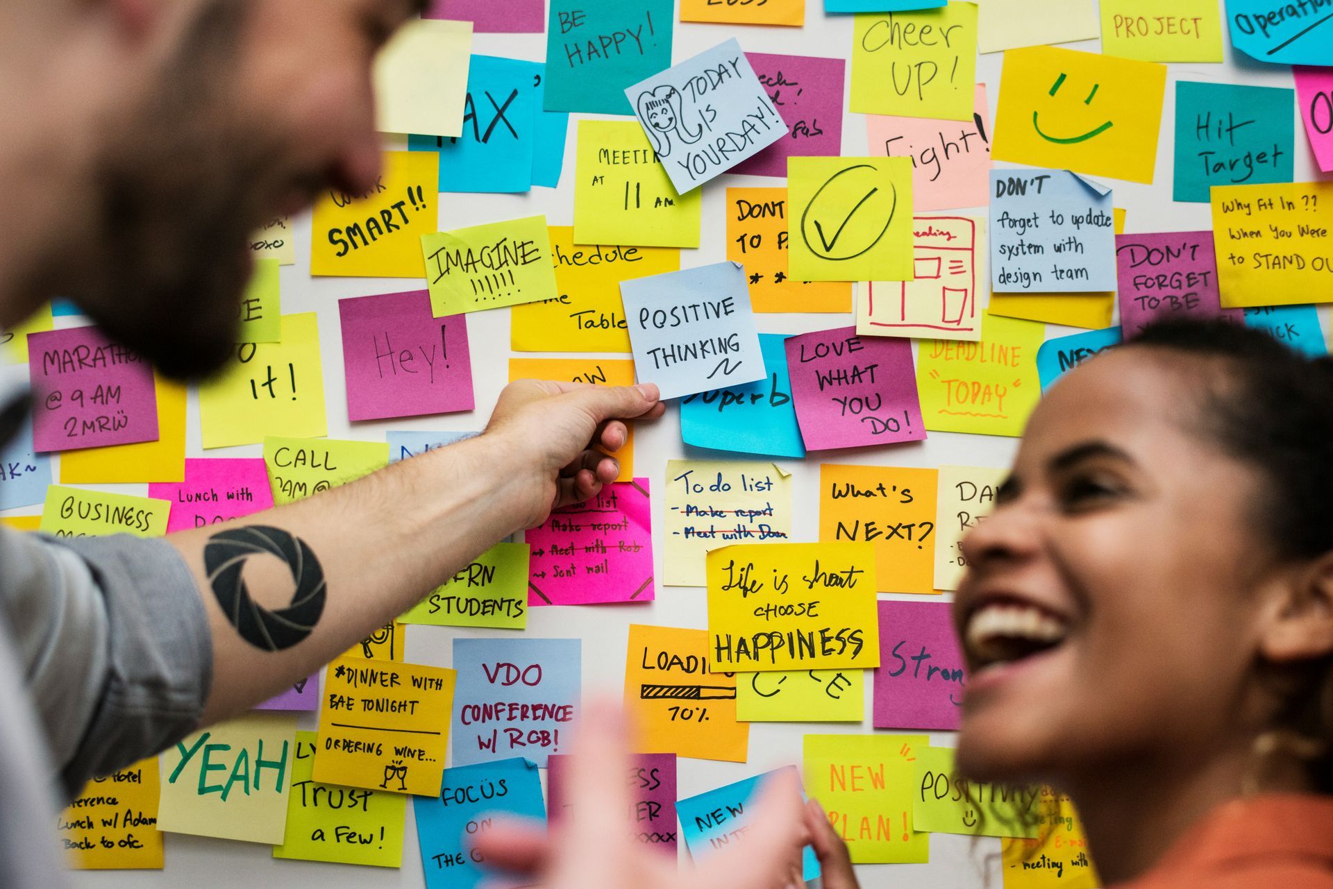 Hand pointing to a 'Positive Thinking' sticky note on a vibrant wall of colorful notes featuring affirming messages like 'Be Happy!', 'Love What You Do', and 'Choose Happiness' - demonstrating emotional expression techniques used in therapy