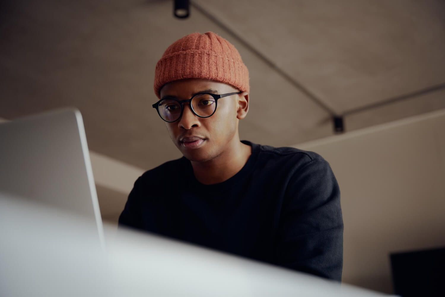 Young Black man wearing glasses and orange beanie looking thoughtfully at laptop screen while rese