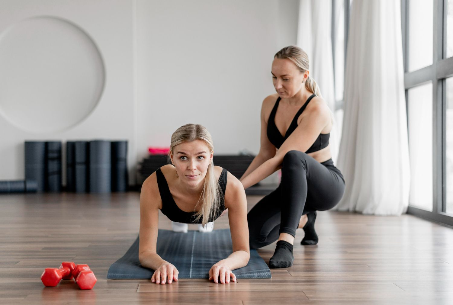 A woman is helping another woman do a plank on a yoga mat.