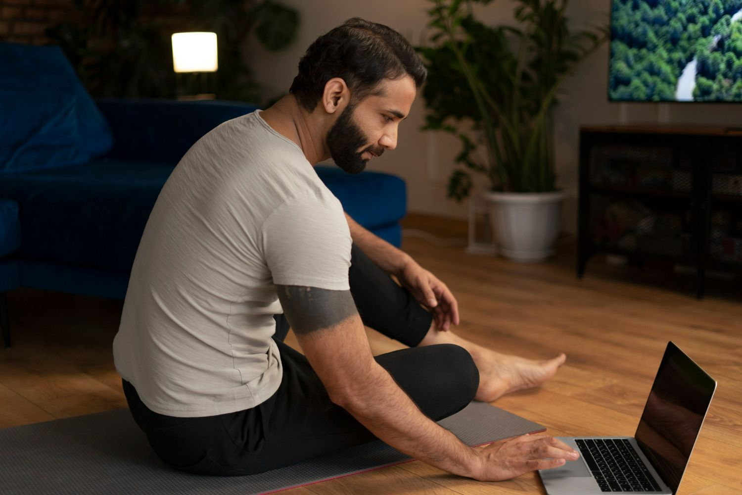 A man is sitting on a yoga mat using a laptop computer.