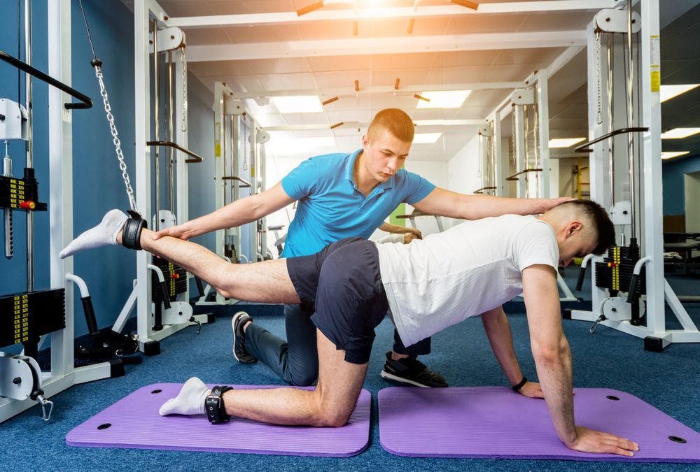 A man is helping another man do exercises on a mat in a gym.