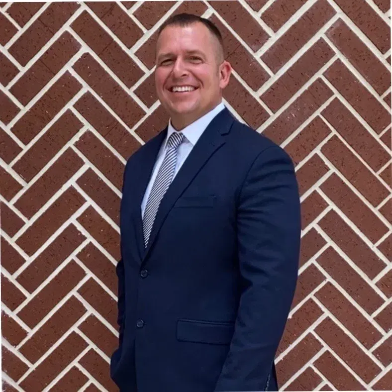 Man in a navy blue suit and tie smiles in front of a brick wall.