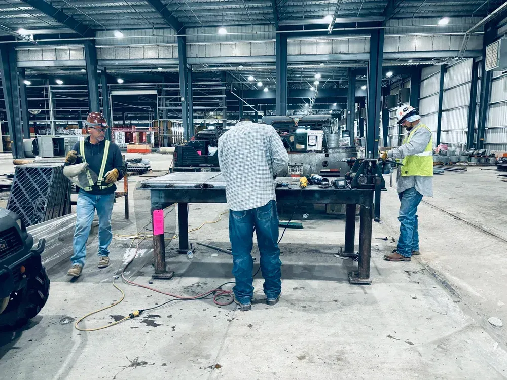 Three construction workers in a metal building, working on a table. One welds.
