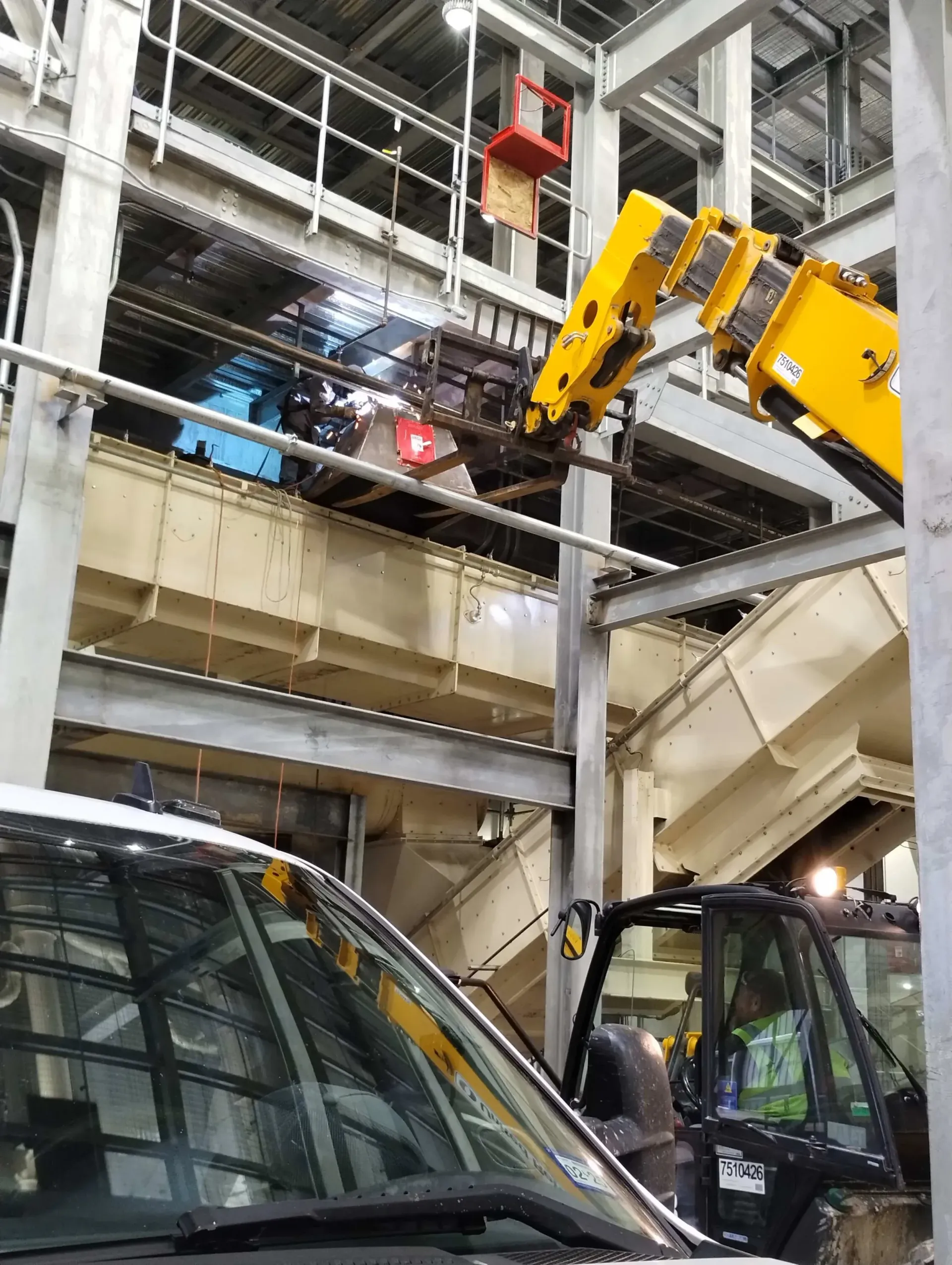 Forklift lifting machinery inside a factory, metal beams and scaffolding visible.