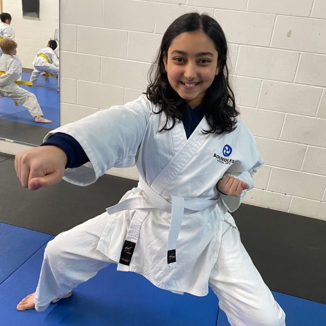 Family in Jiu-Jitsu gis poses. Indoors, blue mats, a smiling woman in the middle, around a seated man with a beard.
