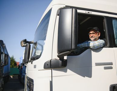 Smiling older truck driver in a white cab, resting arm on the window, looking out at sunny day.