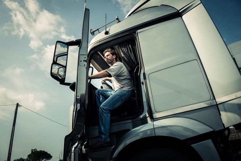 Truck driver exiting the cab, looking towards the viewer, under a bright sky.