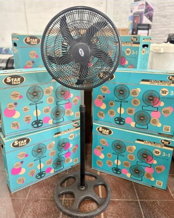 Ventilador de pie negro delante de cajas verde azulado de ventiladores idénticos, en el interior.