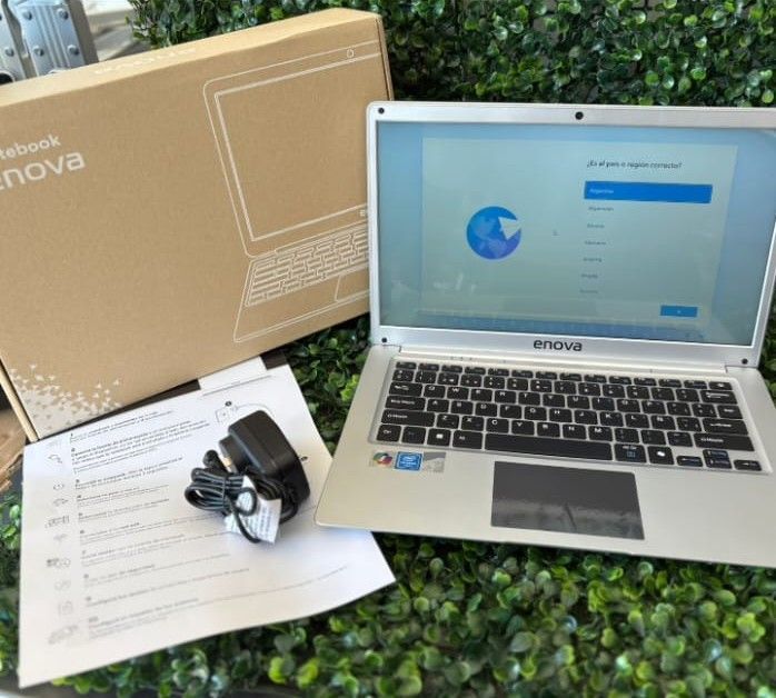 Portátil plateado, cargador y caja sobre follaje verde. La pantalla muestra la configuración de Google.