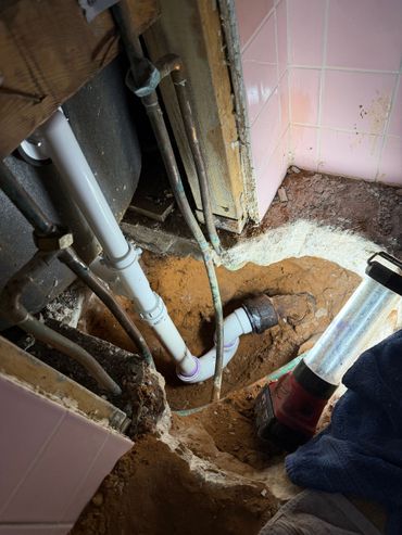 White plumbing pipes installed in a hole in the dirt floor of a bathroom with pink tiles.
