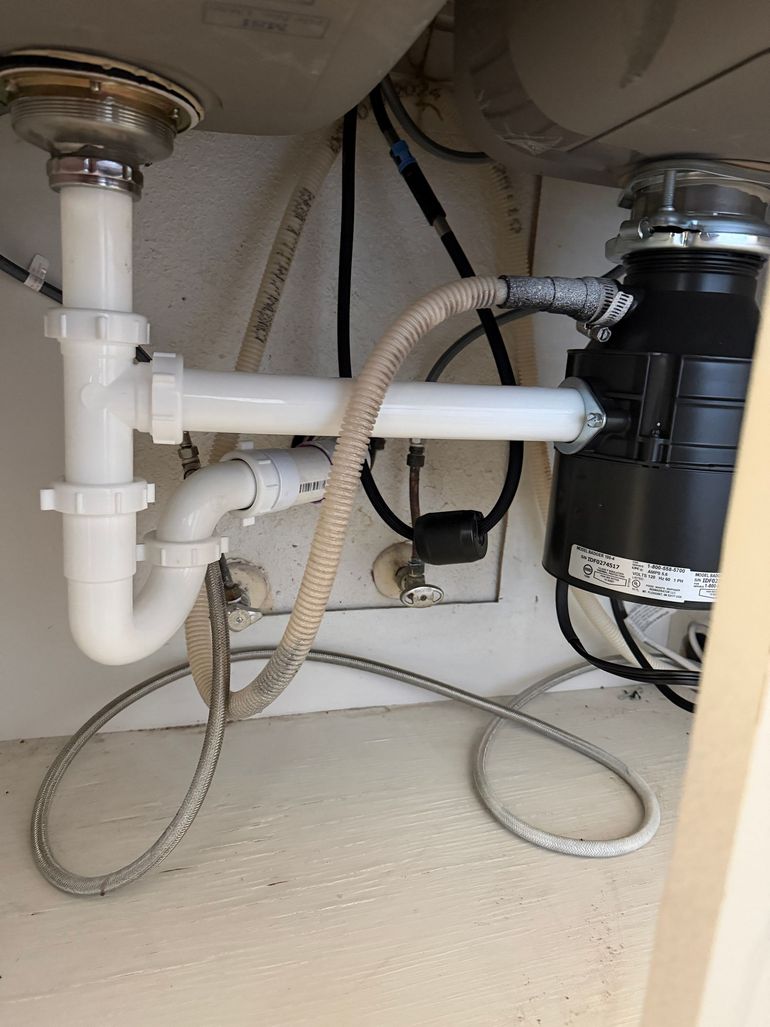 Under-sink plumbing showing a white PVC drain pipe, a p-trap, and a black garbage disposal unit with connected tubing.