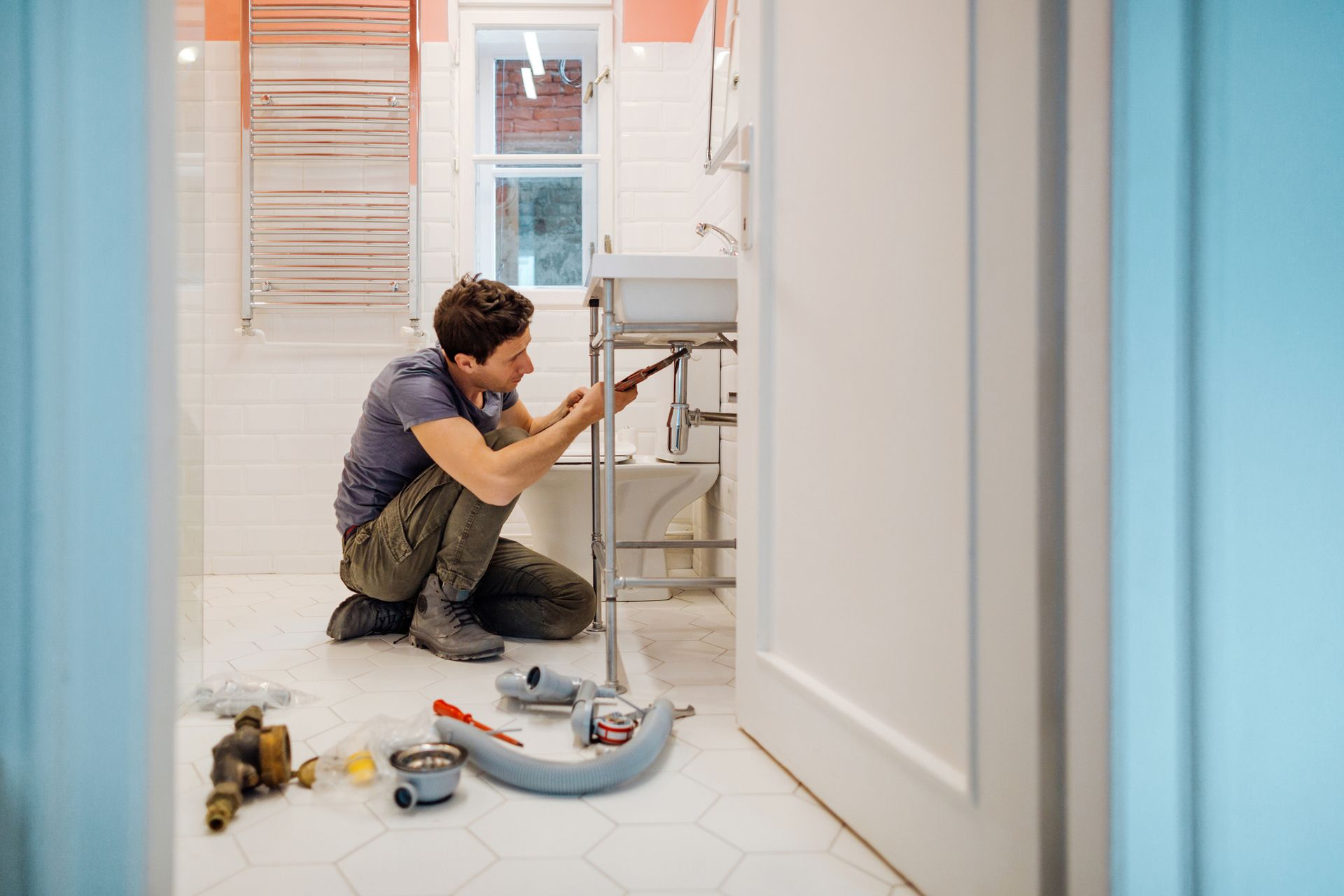 Man kneeling, repairing plumbing under a bathroom sink. Tools scattered on floor.