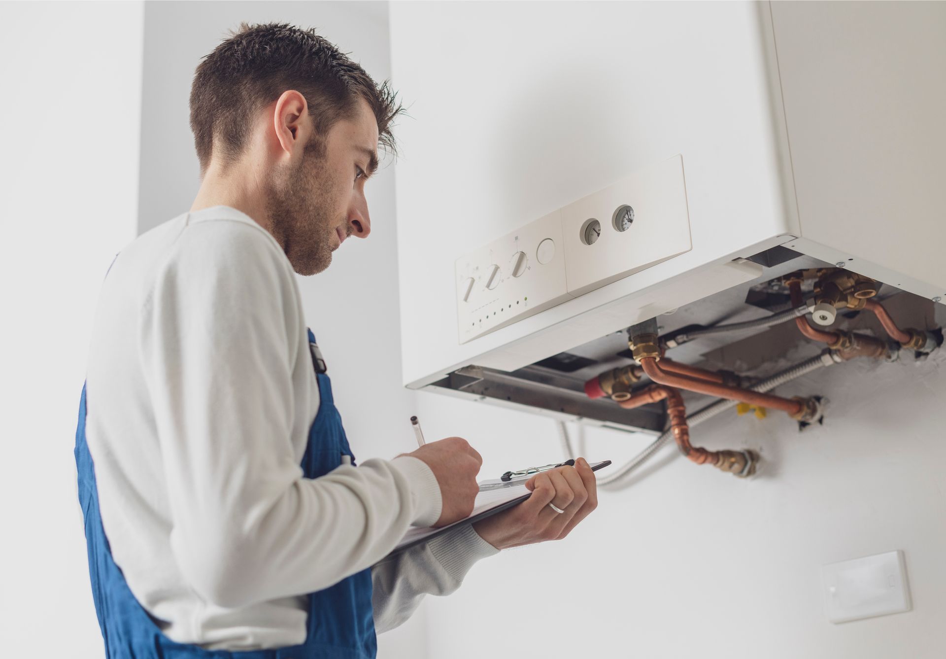 A man in work overalls examines a white wall-mounted boiler, holding a clipboard and pen, indoors.