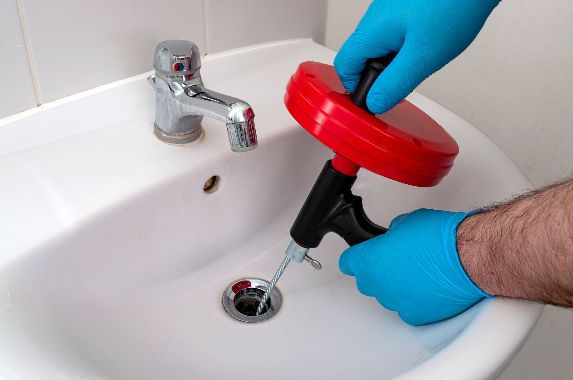 Hands in blue gloves using a drain snake to unclog a white sink in a bathroom.
