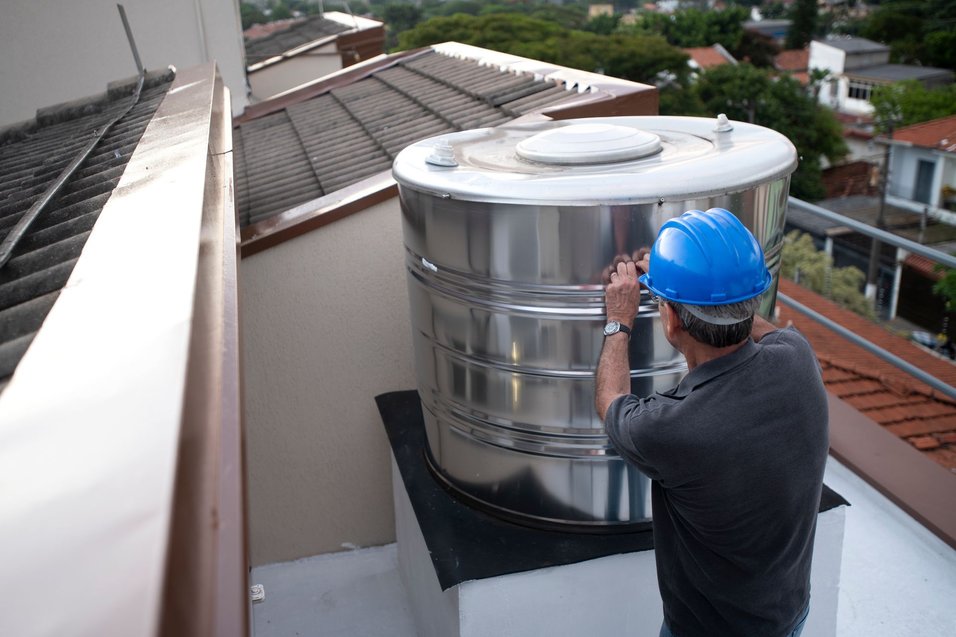 Man in a blue helmet inspects a large stainless steel water tank on a rooftop.