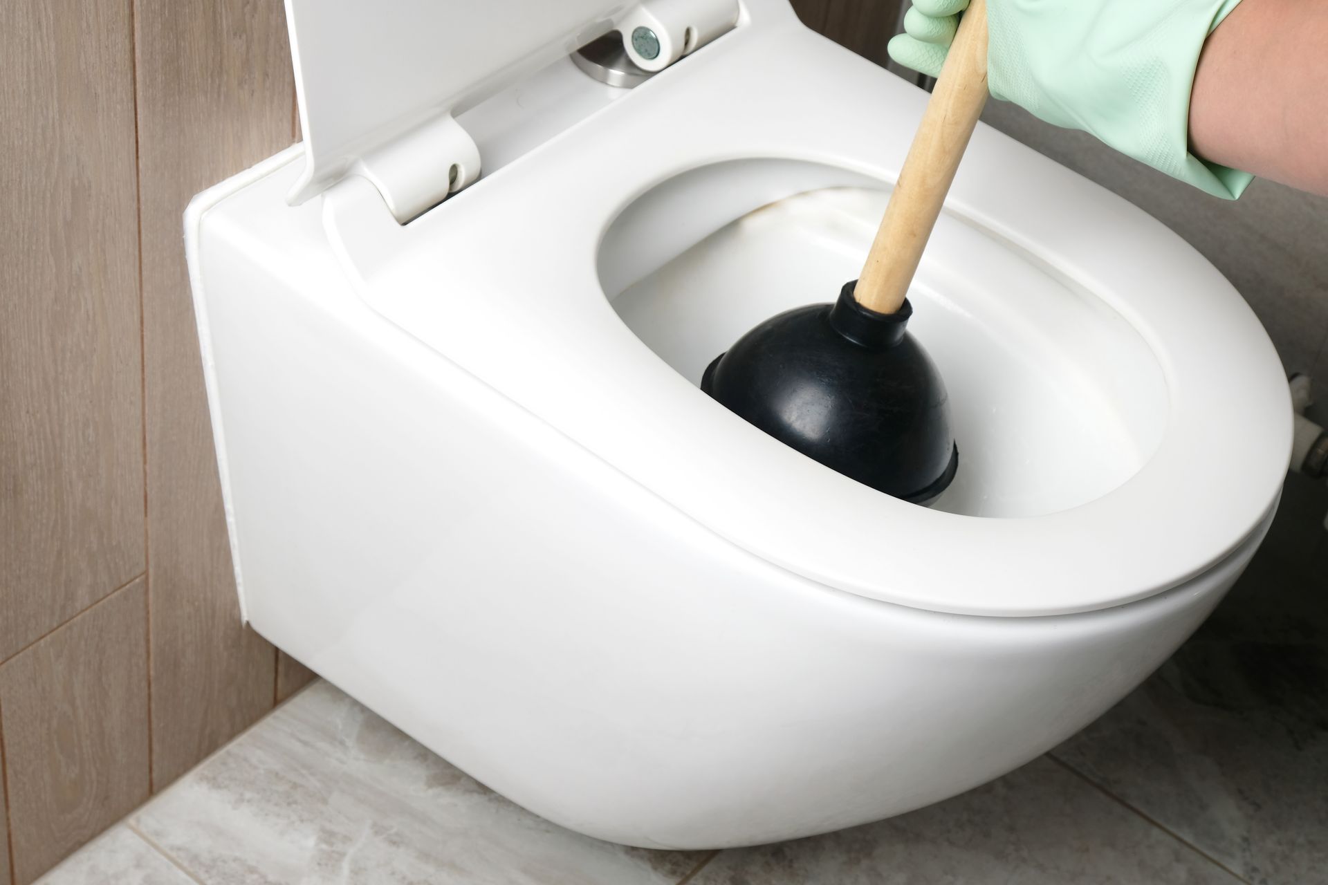 Person in green glove plunges a clogged white toilet in a bathroom.