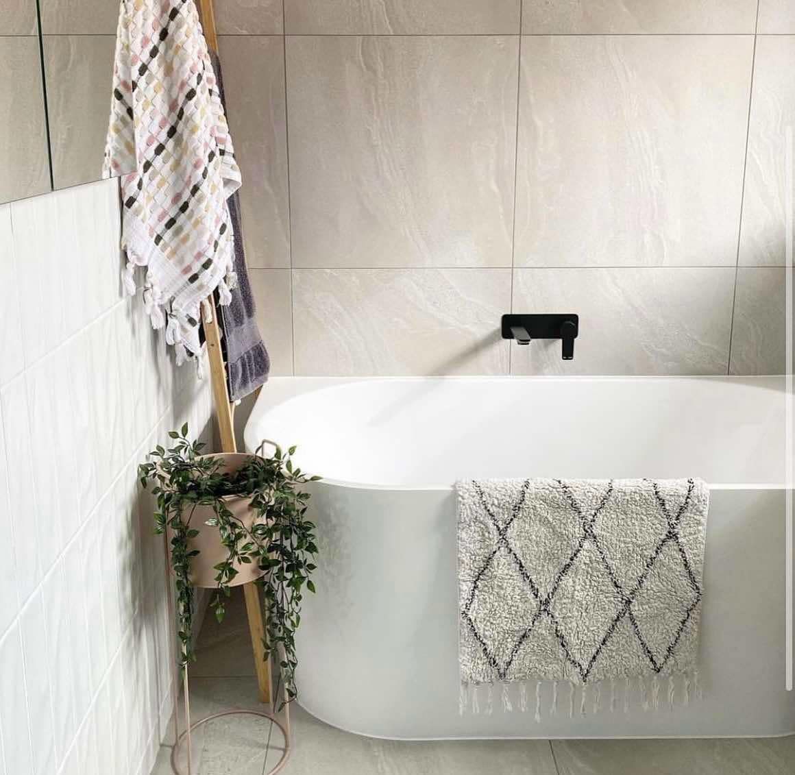 Oval white bathtub with a rug and plant; towels hang on a ladder. Modern bathroom with pale gray walls.
