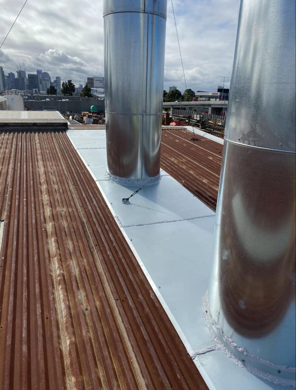 Corrugated metal roof with two silver ventilation pipes; city skyline in background.